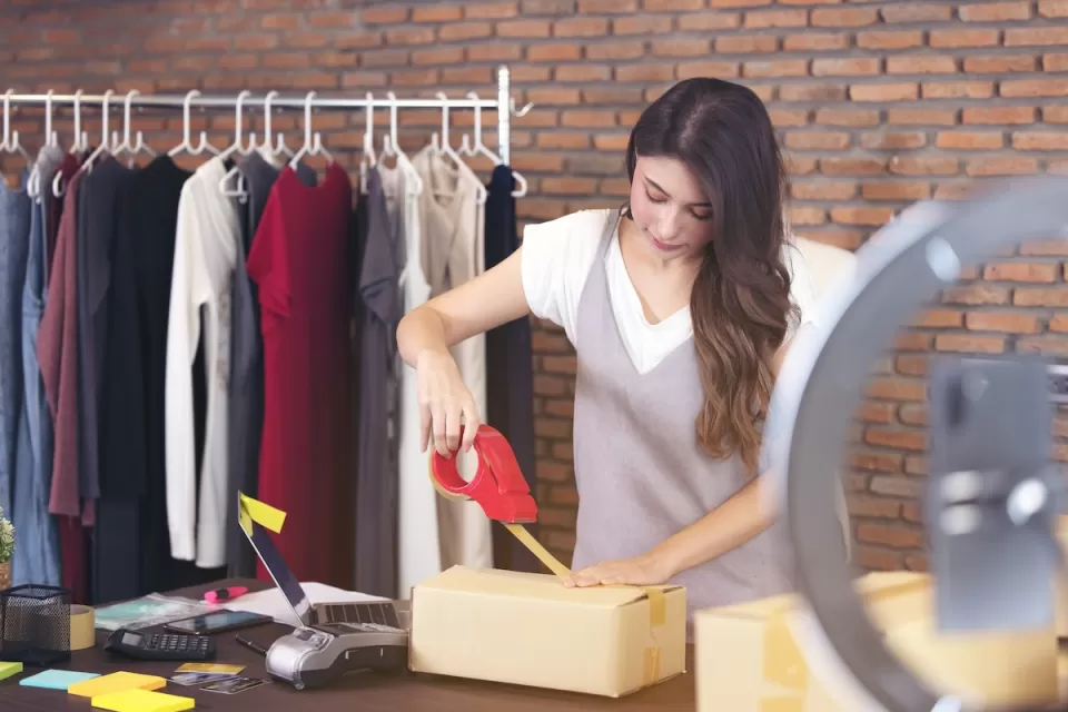 Woman taping up a brown paper package with rack of clothing in background.